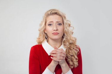 Charming blond woman making capricious face expression and holding hands together in request asking for forgiveness, praying begging on gray. Caucasian person in red formal dress long blond curly hair
