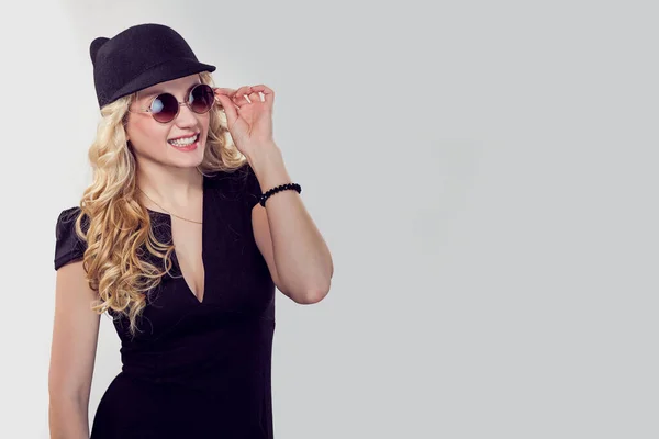 Side view of trendy blond woman in black t-shirt and cap wearing sunglasses and posing smiling flirty with toothy smile on gray. Caucasian Business person in black formal dress, long blond curly hair