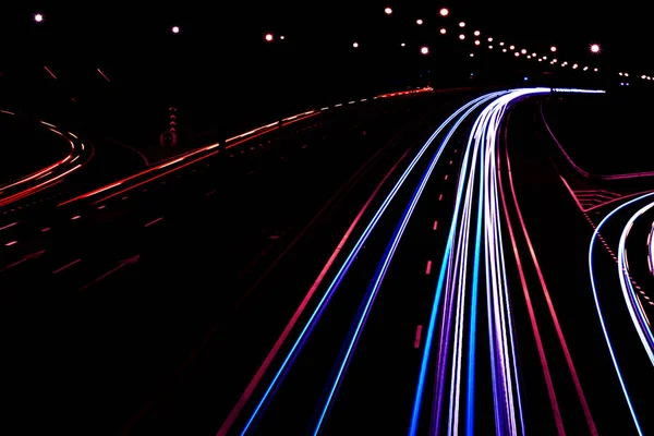 Coches senderos ligeros en una carretera curva por la noche. Rutas de tráfico nocturno. Desenfoque de movimiento. Calle de la ciudad nocturna con movimiento de los faros. Paisaje urbano. Iluminar carretera por vehículo desenfoque de movimiento . — Foto de Stock