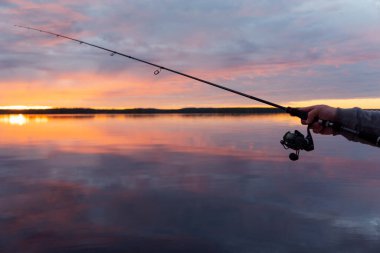 Gün batımında gölde balık tutmak. Elinde olta olan balıkçıların elleri. Makro atış. Balıkçının elleri ve oltası göl suyunun üzerinde. Döndürme çubuğu. Balıkçılık malzemesi
