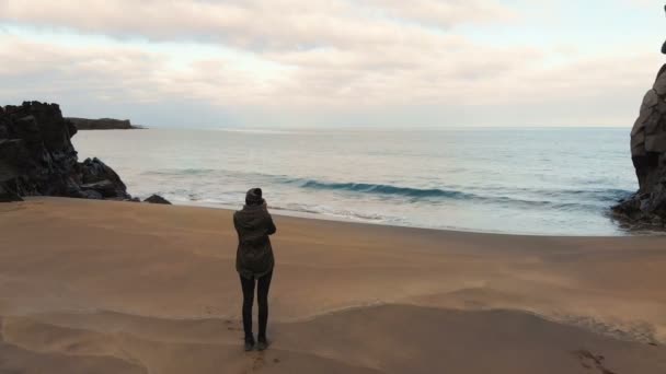 ジャケット姿の若い女性が海を一人で歩き 写真を撮る アイスランドでのコンセプトアドベンチャー スローモーションで空中撮影 ロイヤリティフリーストック映像
