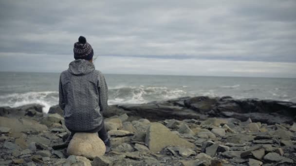 暖かい服の少女は大西洋の崖の海岸に座っている 波が砕けるのを賞賛し ストック映像