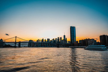 Manhattan ufuk ve nehir üzerinde güzel bir günbatımı. Güneş Manhattan siluetinin arkasında batıyor, nehir kenarındaki köprüleri ve binaları parlak renklerle aydınlatıyor..