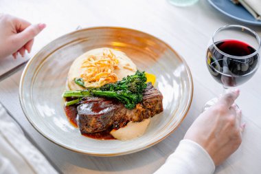 Sofrada gurme yemeği ve şarap var. Biftek, sebze ve kremalı sostan oluşan tabağın keyfini çıkarırken bir el bir bardak kırmızı şarap tutar..