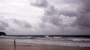Karon sahilinde gün batımı. Denizin üzerinde ağır bulutlar var. Sörf kıyıya vuruyor. Phuket, Tayland