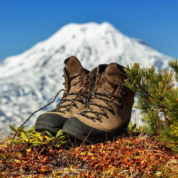 Old trekking boots stand in tundra against background of beautiful cone of volcano on clear sunny day with blue cloudless sky.