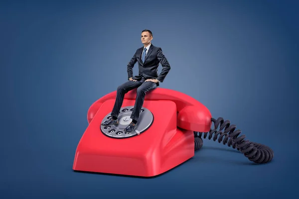 Um pequeno homem de negócios sério senta-se pacientemente em um telefone retro vermelho gigante com um mostrador . — Fotografia de Stock