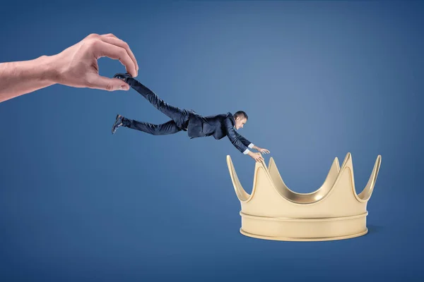 Um pequeno empresário tenta se apossar de uma grande coroa dourada enquanto é arrastado por uma mão enorme . — Fotografia de Stock