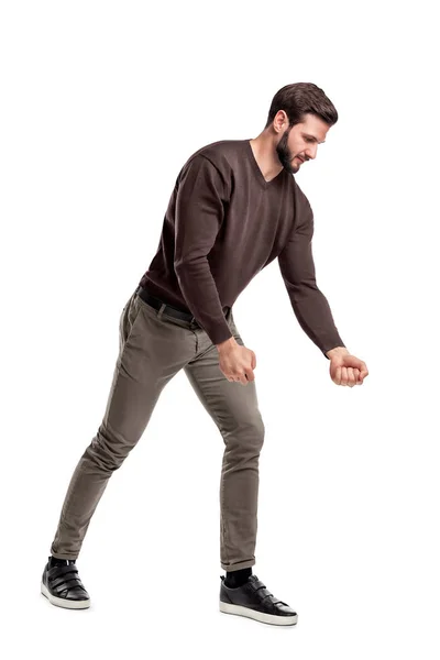 A fit bearded young man in casual clothes tries to pull an invisible rope with all his strength from low grip. — Stock Photo, Image