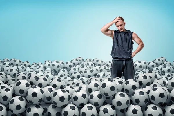 A troubled athletic man stands inside a huge heap of identical football balls.