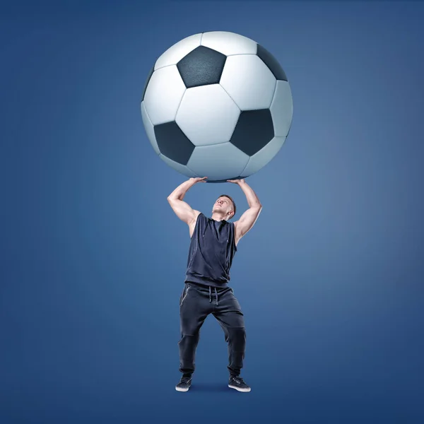 Un hombre musculoso trata de sostener una pelota de fútbol gigante y pesada en blanco y negro sobre sí mismo . — Foto de Stock