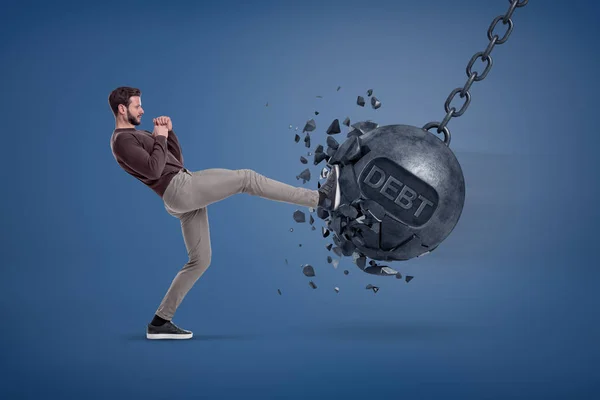 En man i casual kläder sparkar en giant wrecking ball med ett ord skuld skrivet på det. — Stockfoto