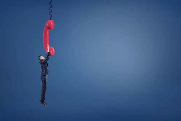 Een kleine zakenman heeft een gigantische rode retro telefoon handvat en hangt aan het proberen niet te vallen. — Stockfoto