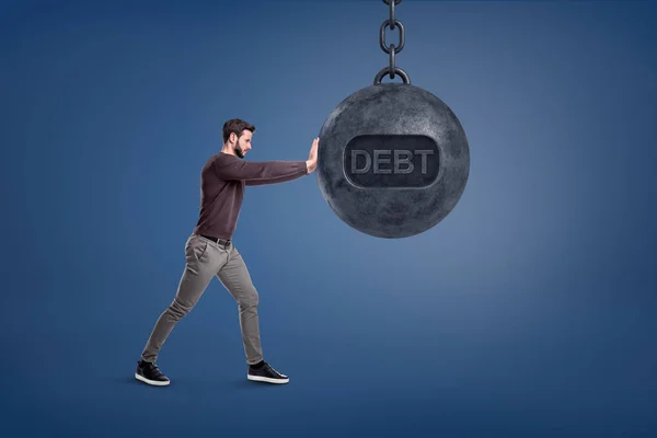 Um homem barbudo está perto de uma enorme bola de demolição com uma palavra Dívida nele e tenta empurrá-lo . — Fotografia de Stock