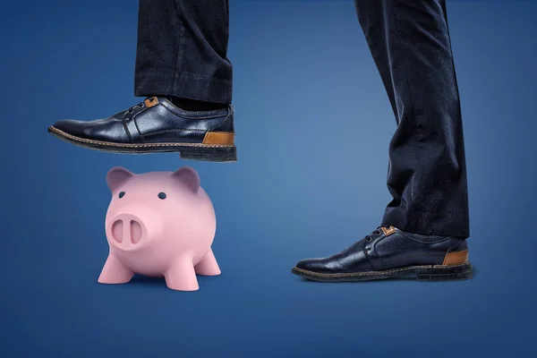 Een roze spaarvarken staat onder een gevaarlijke mannelijke voet die klaar zijn om stap op het. — Stockfoto