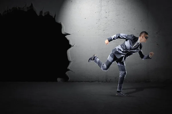 Un jeune homme d'affaires en costume rayé et un masque noir fuyant un trou noir dans un mur . — Photo