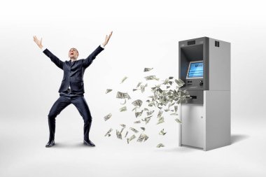 Etrafta dolaşan birçok doları banknot ile bir ATM yakınındaki beyaz bir arka plan üzerinde mutlu bir işadamı duruyor.