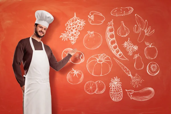 Un buen cocinero joven en un sombrero de chef y delantal haciendo un gesto de presentación a una imagen de frutas y verduras en una pared . —  Fotos de Stock