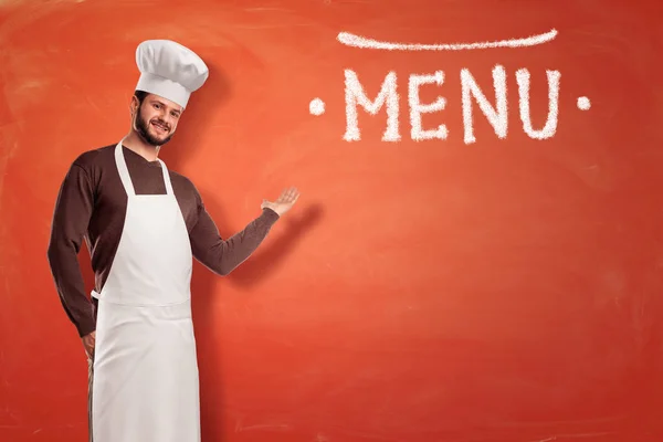 Brodaty kucharz w biały uniform stoi i punkty do wskazania menu dużych kreda rysowane. — Zdjęcie stockowe