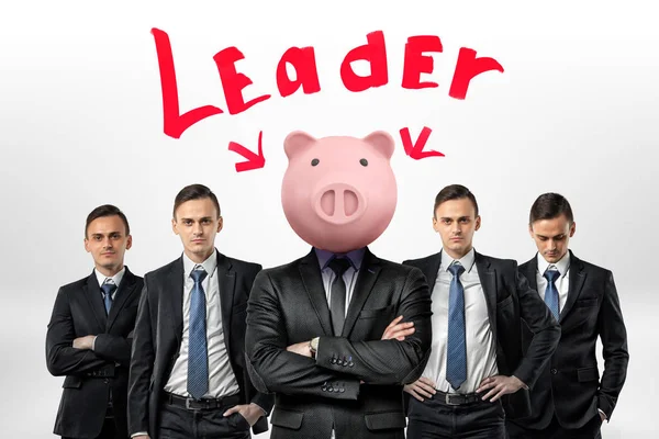 Varios hombres de negocios idénticos se visten entre sí alrededor de un hombre con una cabeza de alcancía bajo la palabra Líder. T — Foto de Stock
