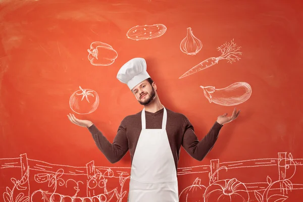 Un buen chef joven en un sombrero de chef y delantal haciendo un gesto de presentación a las imágenes de verduras en una pared . —  Fotos de Stock