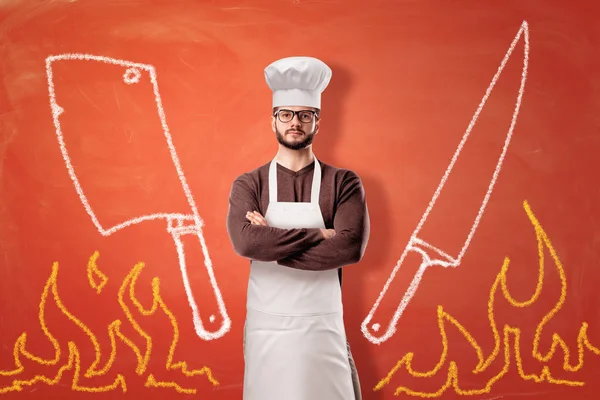 Un fondo naranja brillante con llamas dibujadas, una cuchilla, un cuchillo y un cocinero masculino serio de pie en el centro . —  Fotos de Stock