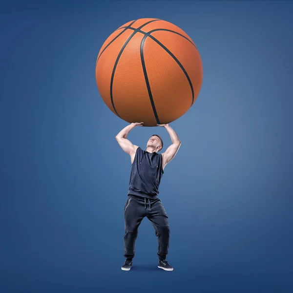 Un joven musculoso sostiene una pelota de baloncesto naranja gigante sobre su cabeza. . — Foto de Stock