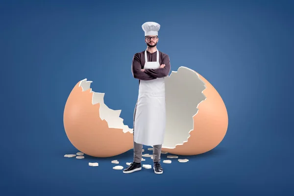 Un joven cocinero barbudo en un delantal blanco y un sombrero se encuentra cerca de una cáscara de huevo rota grande . —  Fotos de Stock