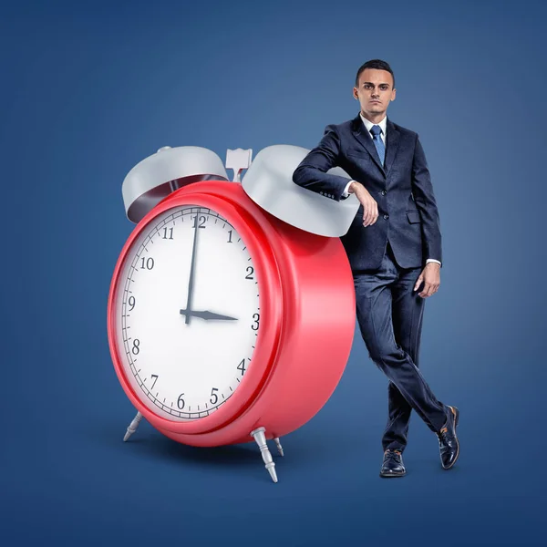 A small businessman stands looking at front and leaning on a huge red alarm clock. — Stock Photo, Image