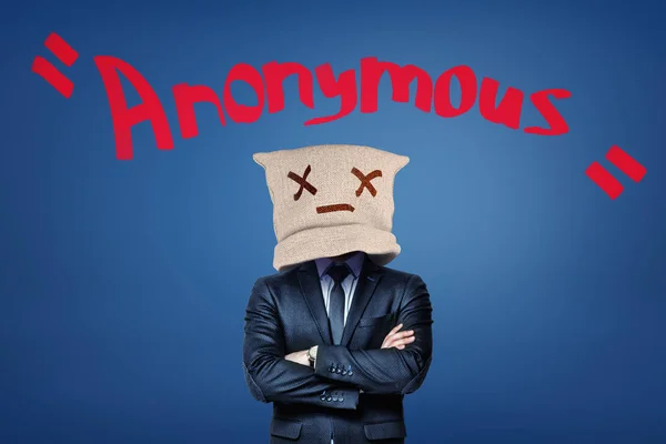 A man in suit with a canvas bag on his head with a dead emoticon on it, standing with arms folded, with the title Anonymous above.