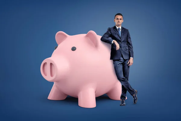 Ein junger Geschäftsmann steht neben einem großen rosafarbenen Sparschwein und lehnt daran. — Stockfoto