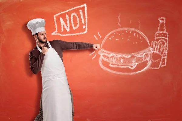 Un joven chef guapo diciendo No y golpeando con los puños cerrados un dibujo de tiza de una hamburguesa, papas fritas y una botella de ketchup . —  Fotos de Stock