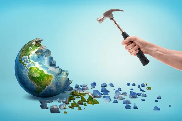 Een mannenhand houdt een metalen hamer in de buurt van een semi-gebroken Earth globe met kleine stukjes uit het geraakt. — Stockfoto