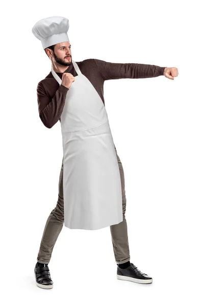 Un joven y apuesto chef con sombrero de chef y delantal posando con los puños cerrados y la mano izquierda extendida como si estuviera a punto de golpear algo . —  Fotos de Stock