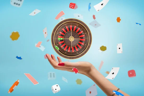 Female hand with a roulette, white dice, playing cards and stacks of chips in the air on blue background
