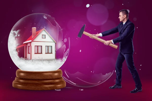 Egy üzletember egy kalapáccsal a kezében összeomlik a kristálygömb, egy kis ház belsejében egy lila háttér, néhány pöttyök a hó. — Stock Fotó