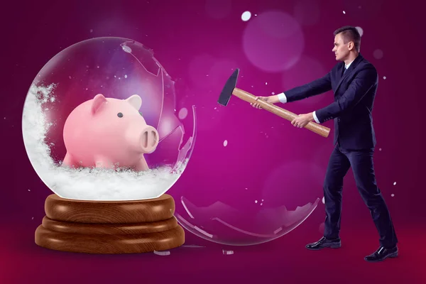 Un homme d'affaires avec un marteau dans les mains écrasant une grosse boule de cristal avec une tirelire à l'intérieur sur un fond violet avec quelques éclats de neige . — Photo