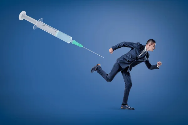 A side view of a businessman running from a huge syringe floating in the air on a blue background.
