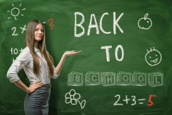 Uma mulher de negócios bonita ou um professor de pé meia volta em um fundo da diretoria da escola apontando para o título Back to School nele . — Fotografia de Stock