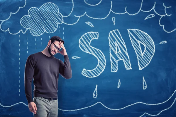 Young man with SAD chalk sign and pouring rain clouds drawn on blue board background