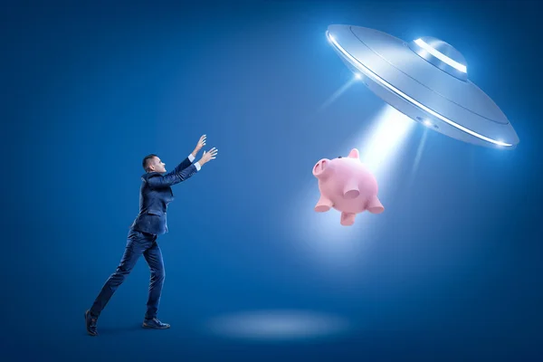 Ein Geschäftsmann hebt die Hände zu einem Ufo, das ein Sparschwein durch eine unsichtbare Kraft zu seiner offenen Luke zieht. — Stockfoto