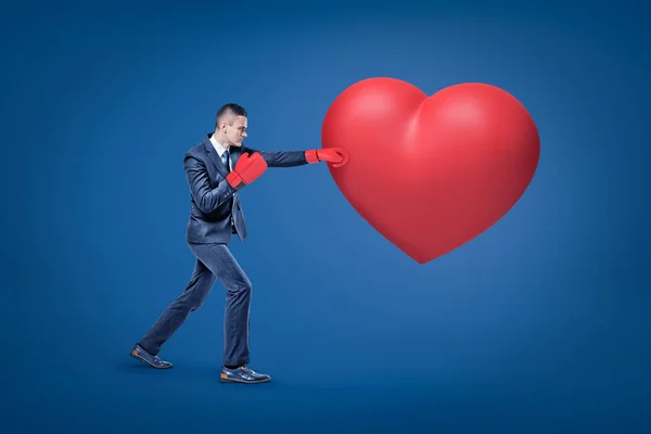 Businessman with red boxing gloves punching big red heart on blue background