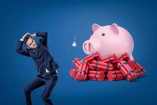 Zakenman beschermen zichzelf met handen en roze spaarvarken over Rode dynamiet stokken met verlichte zekering op blauwe achtergrond — Stockfoto