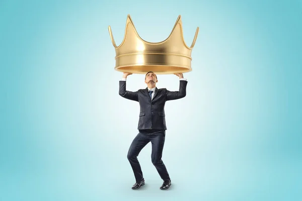 Jovem empresário segurando heady coroa dourada acima da cabeça no fundo azul — Fotografia de Stock