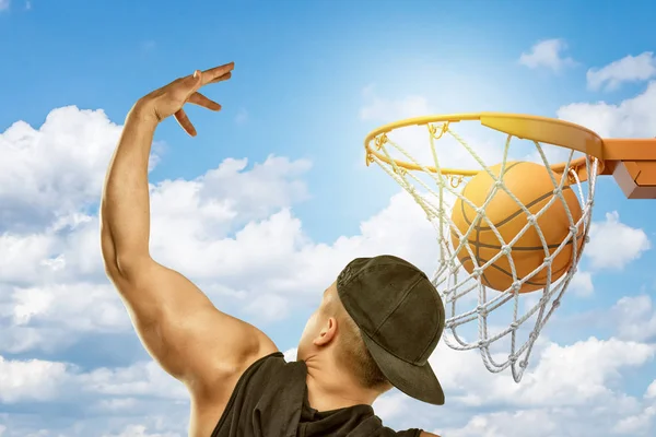 Hombre vistiendo camiseta negra y gorra negra lanzando pelota de baloncesto en aro sobre fondo de nubes blancas cielo azul — Foto de Stock