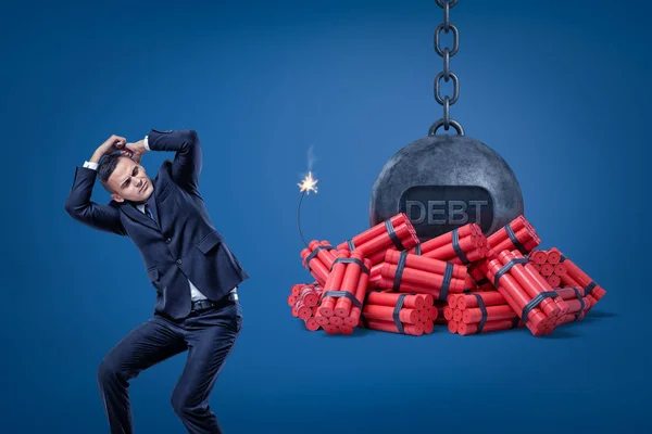 Empresario protegiéndose con las manos y bola encadenada con un signo DEBT sobre palos de dinamita roja con fusible encendido sobre fondo azul — Foto de Stock
