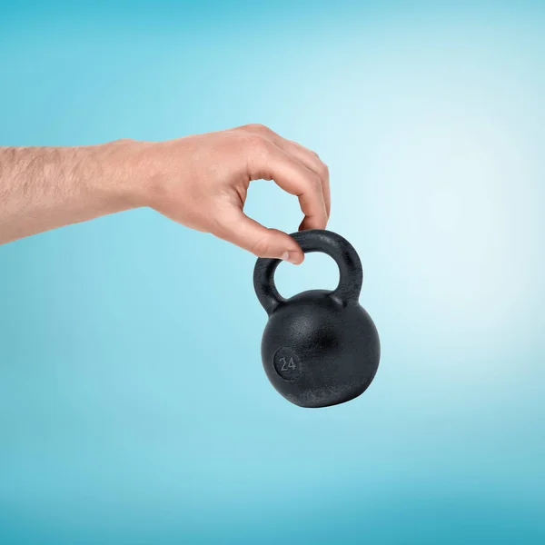 Male hand holding small black kettlebell between two fingers on blue background — Stock Photo, Image