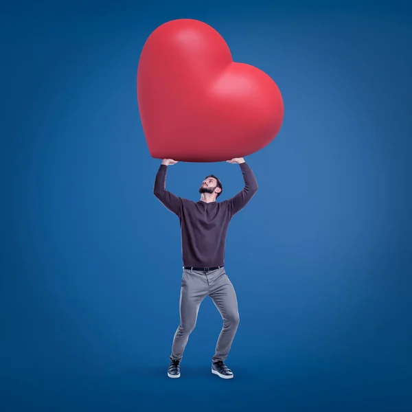 Joven hombre en forma en ropa casual sosteniendo enorme corazón rojo de San Valentín sobre fondo azul . — Foto de Stock