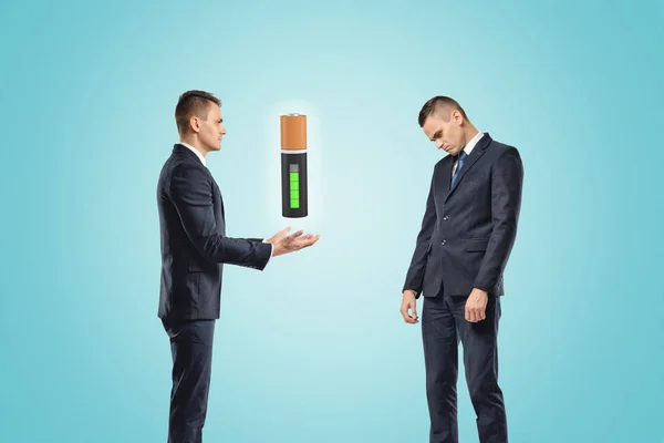 Two businessmen, one posing as if manually controlling the battery suspended in the air, the other standing half-turned with a sad expression on his face.