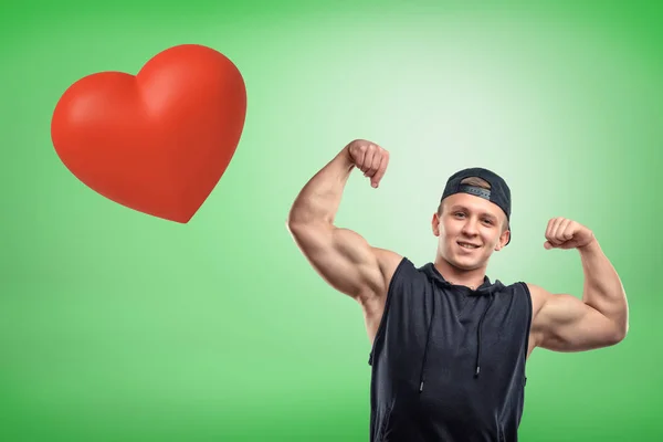 Starker muskulöser junger Mann zeigt Bizeps mit großem roten Herz auf grünem Hintergrund — Stockfoto